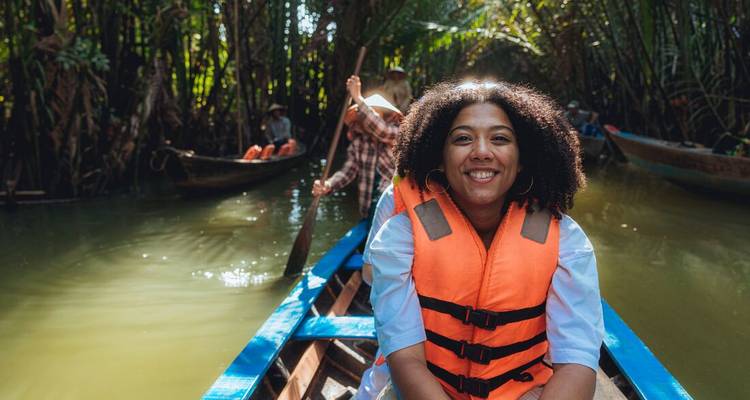 Een glimlachende vrouw in een oranje reddingsvest zit in een houten sampan die voortglijdt door een schaduwrijke kanaal in het Mekongdelta.