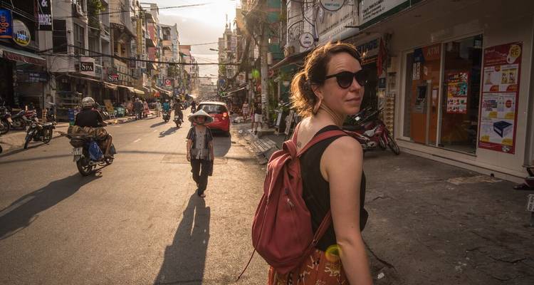 Een reiziger met rugzak kijkt terug langs een zonnige Vietnamese straat vol met winkels en scooters.