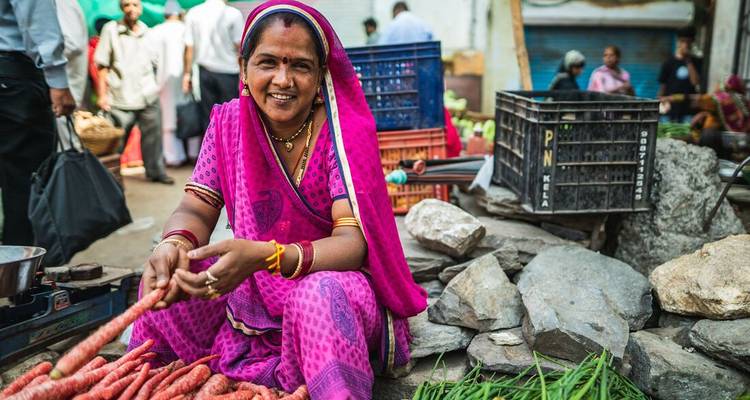 Mujer con un vestido tradicional vibrante vendiendo verduras en un mercado al aire libre.