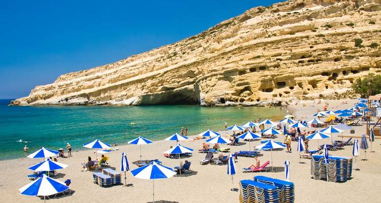 Belebter Sandstrand mit blau-weißen Sonnenschirmen unterhalb dramatischer geschichteter Klippen und türkisfarbenem Meer