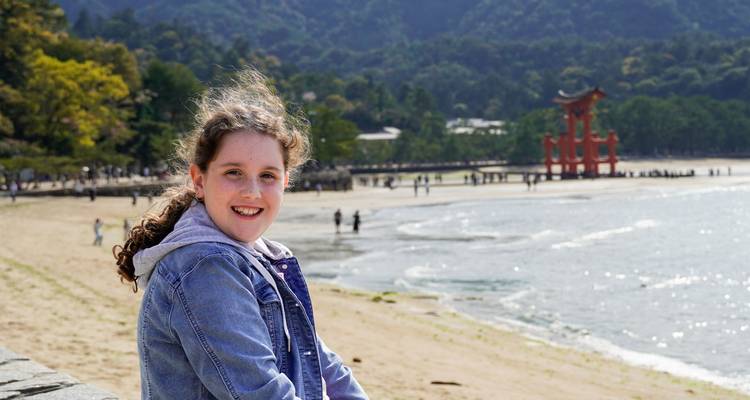 Una chica sonríe en la playa arenosa de Miyajima con la famosa puerta torii roja elevándose desde el mar detrás de ella.