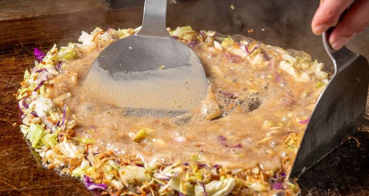 El okonomiyaki chisporrotea en una plancha mientras las espátulas mezclan la masa burbujeante con verduras de colores.