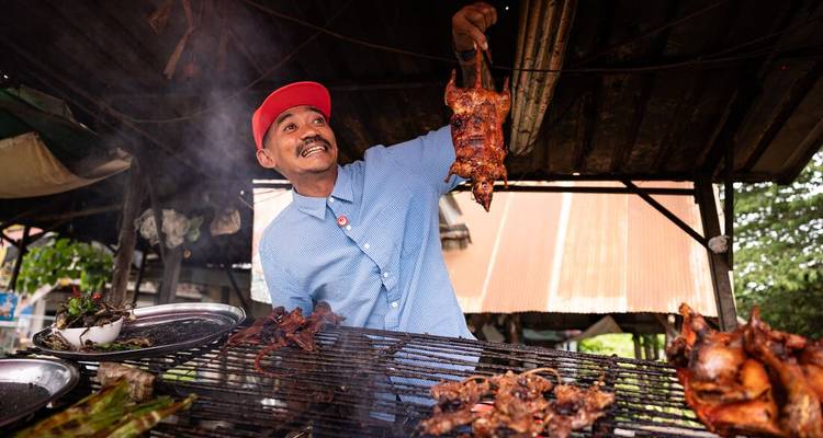 Un vendedor de comida callejera sostiene orgullosamente un animal asado en medio de parrillas humeantes y brochetas.