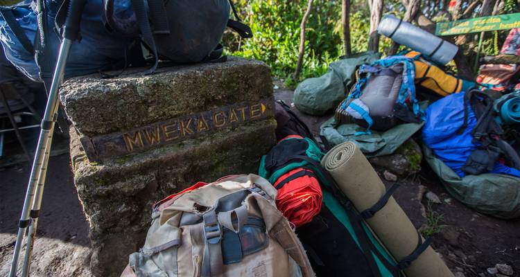 Les sacs à dos s'empilent à côté du marqueur en pierre indiquant « Mweka Gate » à l'entrée du sentier forestier.