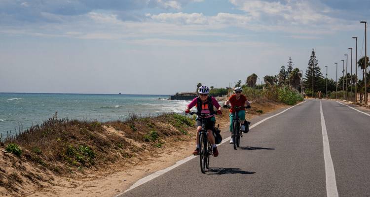 Dos ciclistas andan en bicicleta por una carretera costera con el Mar Mediterráneo y algunas palmeras a su izquierda.
