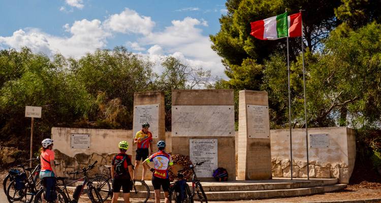 Los ciclistas se detienen en un monumento rural que tiene la bandera italiana ondulando, escuchando a su guía.