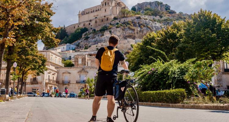 Un ciclista con una mochila amarilla contempla la catedral barroca de Modica elevándose sobre el pueblo en la ladera.
