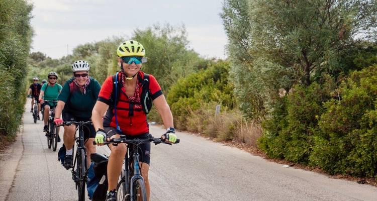 Una fila de ciclistas recorre un estrecho camino rural rodeado de matorrales mediterráneos densos.