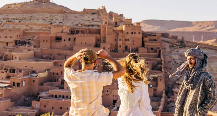 Reisende und ein einheimischer Mann mit Turban bewundern die Lehmkasbahs von Ait Benhaddou von einem malerischen Aussichtspunkt bei Sonnenuntergang.