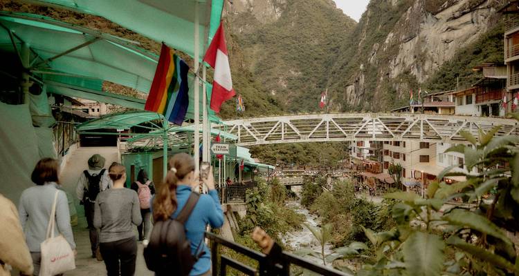 Travelers walk a covered pathway lined with flags toward a pedestrian bridge above a rushing river in a mountain town.