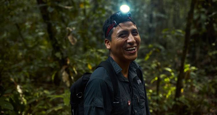 Smiling jungle guide wearing a headlamp stands in dense Amazon rainforest at dusk.