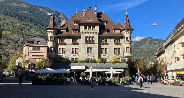 Bustling square in front of an ornate historic building flanked by alpine hills and clear skies.