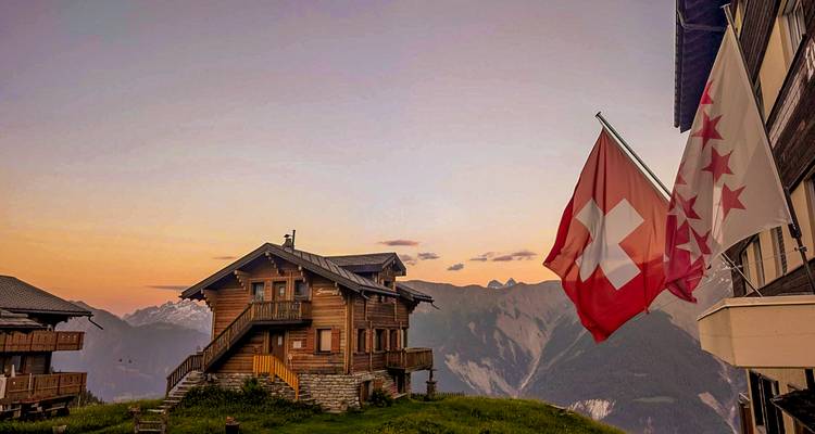 Swiss flags flutter beside a wooden chalet overlooking alpine peaks at warm twilight.