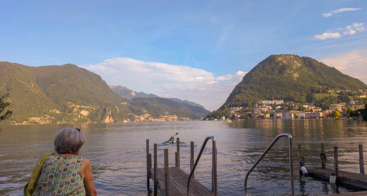 A woman watches a kayaker from a lakeside pier against the verdant slopes surrounding Lake Lugano.