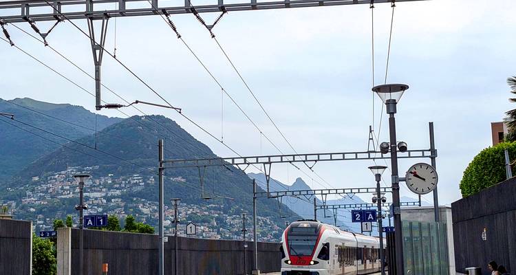 Modern Swiss train at a small station with mountainous backdrop and overhead lines.