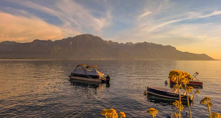 Small boats drift on Lake Geneva at sunset with the dark silhouette of the Alps behind.