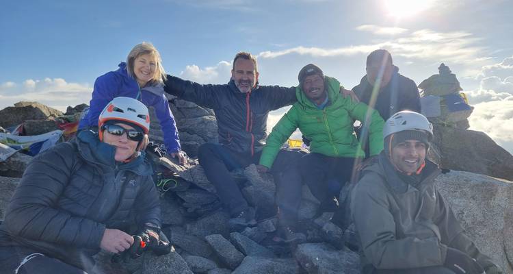 Montañeros con equipo de invierno celebran en una cumbre rocosa con banderas de oración bajo un sol brillante.