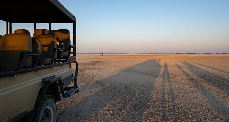 Safari vehicle casts long shadows across the barren white expanse of Botswana’s salt pans at dusk.