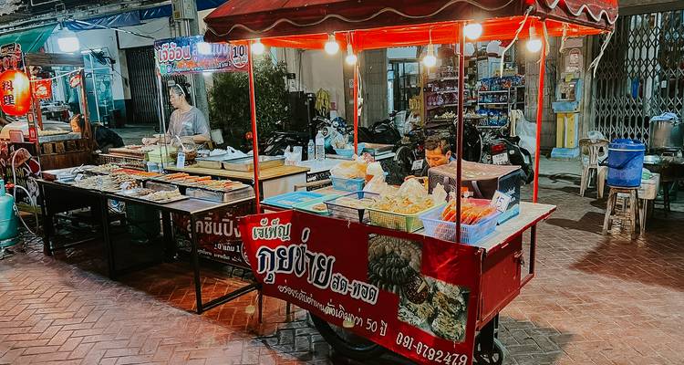 Carrito de comida callejera nocturno en Tailandia brillantemente iluminado con bandejas de snacks y un vendedor preparando pedidos.