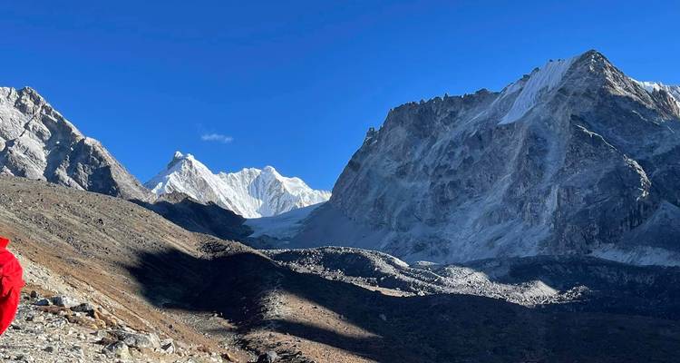 Solo trekker in een rood jasje die langs een rotsachtig Himalaya-pad wandelt met torenhoge sneeuwtoppen vooruit.