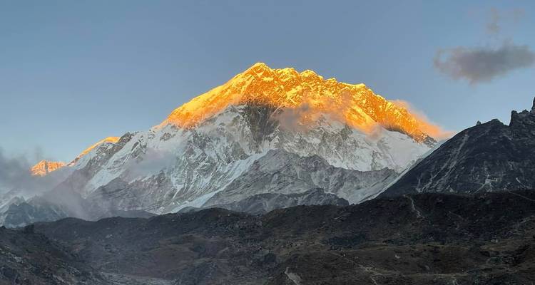 Besneeuwde Himalaya-top die tijdens zonsopgang oranje oplicht, terwijl sliertjes wolk over de bergrug drijven.