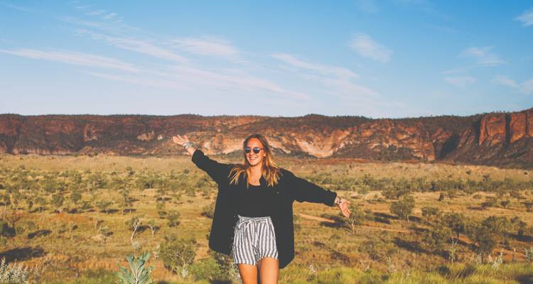 Junge Frau mit weit ausgebreiteten Armen steht in Australiens roter Outback mit Steilküste im Hintergrund.