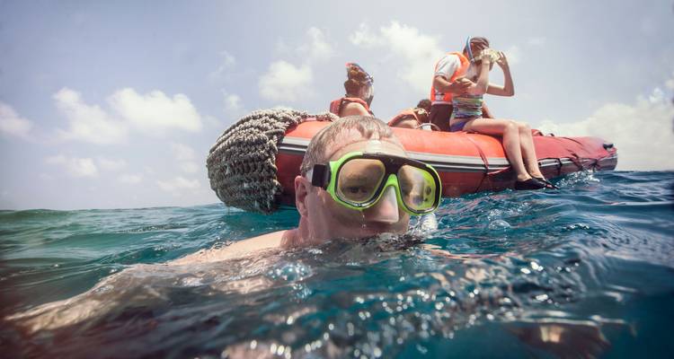 Snorkelaar op de voorgrond met een snorkelmasker terwijl vrienden op een opblaasbare boot zitten onder een helder lucht.