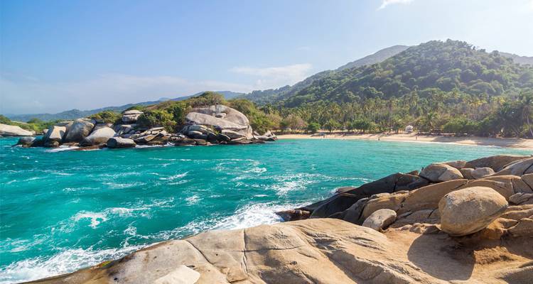 Turquoise Caribbean bay with rocky shoreline, sandy beach and lush mountains in the background.