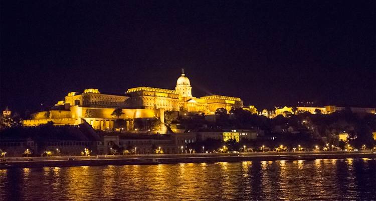 El Castillo de Buda brillando en tono dorado en una noche oscura con reflejos brillantes en el Danubio.