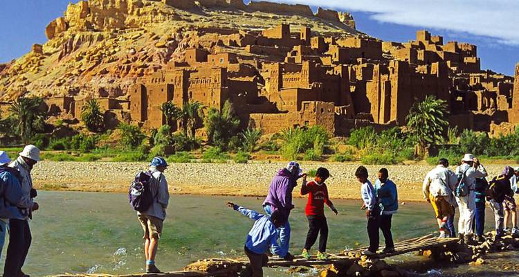 Visitors carefully cross a makeshift footbridge over a shallow river with the mud-brick fortress of Aït Benhaddou rising behind.