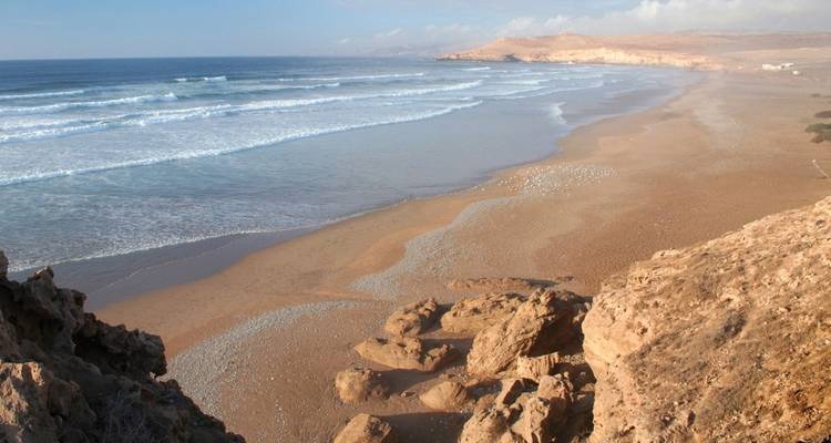 Wide sandy Atlantic beach with gentle waves and rocky cliffs under hazy skies.