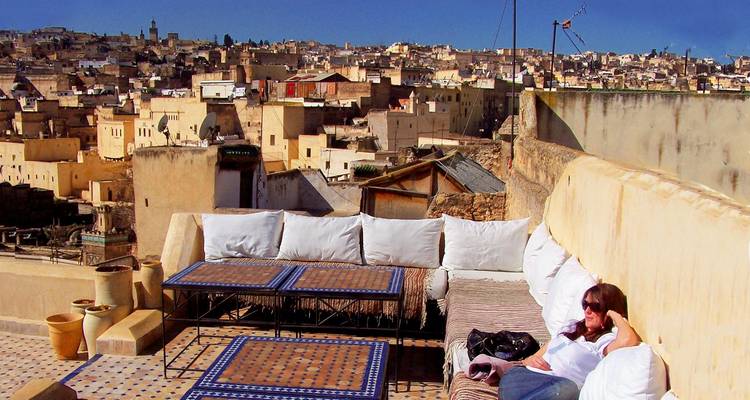 Woman relaxing on a cushioned rooftop terrace overlooking a dense ochre-colored medina of flat-roofed houses under clear blue sky.