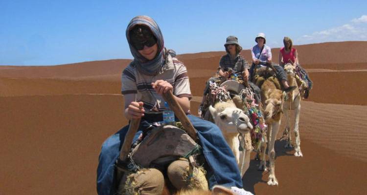 Caravan of travellers riding camels in single file across towering orange Sahara dunes beneath a clear sky.