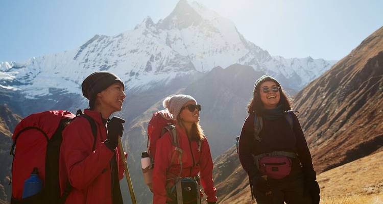 Three trekkers in red jackets smile beneath towering snow-clad Himalayan peak.