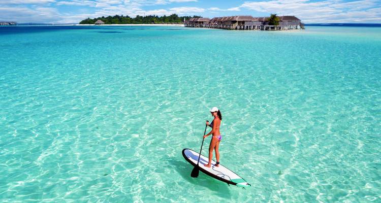 A woman stand-up paddles over crystal-clear turquoise water with over-water villas and a lush island in the background.