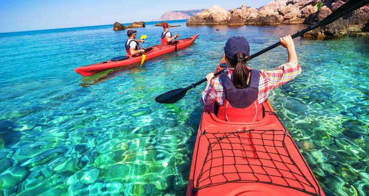 Kayakers paddle bright red boats across sparkling, translucent tropical waters near rocky shores.
