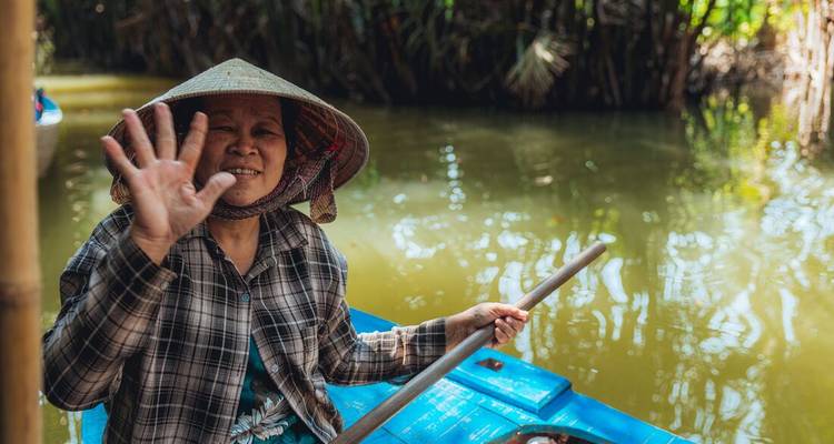 Femme locale souriante agitant la main en ramant dans une barque en bois bleu le long d'un étroit canal de jungle