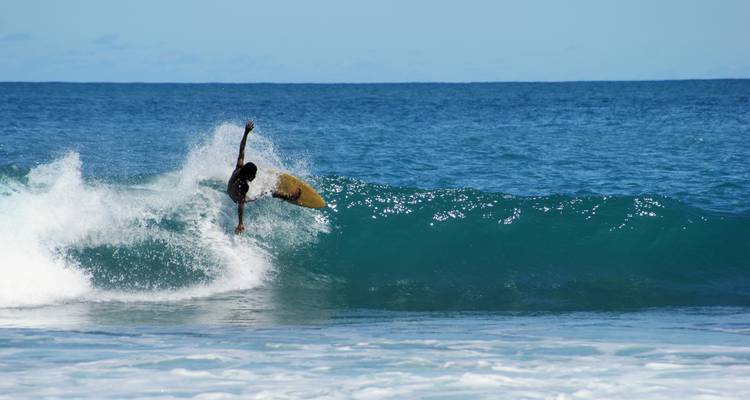 Un surfeur traçant une courbe sur une vague turquoise avec des embruns blancs sous un ciel bleu dégagé.