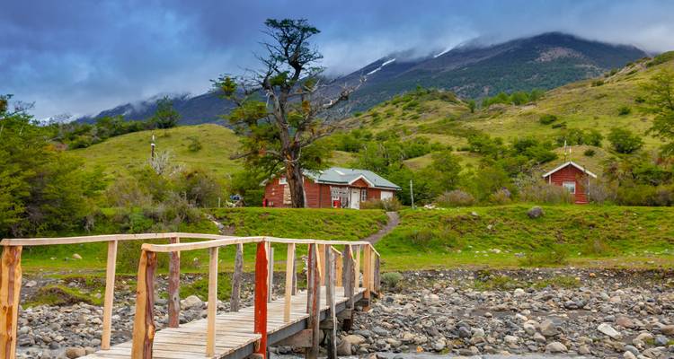 Petit pont en bois menant à des cabanes rustiques sur une colline herbeuse sous les pics des Andes couverts de nuages.