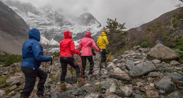 Quatre trekkeurs vêtus de vestes de pluie colorées gravissent un sentier rocheux en direction de montagnes enneigées enveloppées de brume.