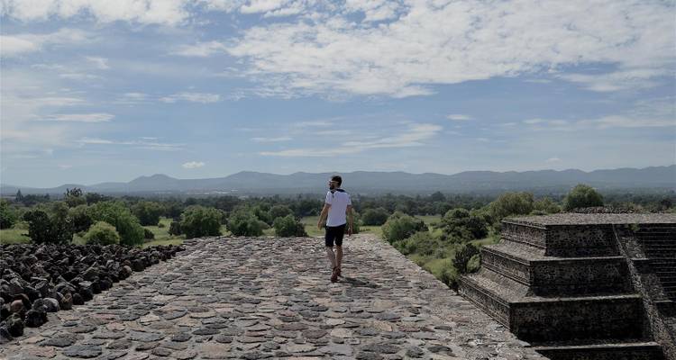 A traveller walks along the stone summit of a pyramid at Teotihuacan overlooking green plains and mountains.