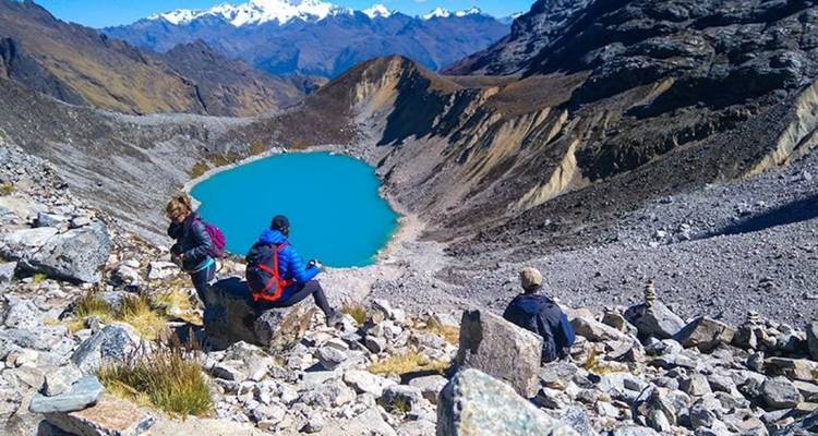 Excursionistas sentados en rocas con vista a un lago azul de montaña y picos nevados.
