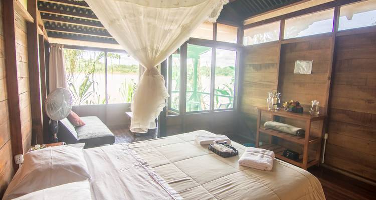 Airy wooden lodge bedroom with mosquito-netted bed, large windows overlooking a river and surrounding jungle.