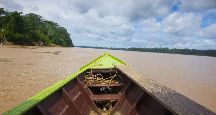 Forward view from a small wooden boat gliding along a wide muddy jungle river under broken clouds.
