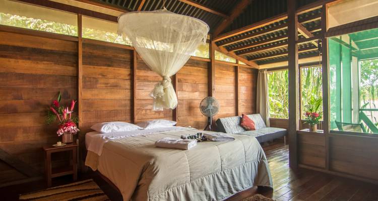 Spacious wooden bedroom with king bed, white mosquito net, fan and jungle views through screened windows.
