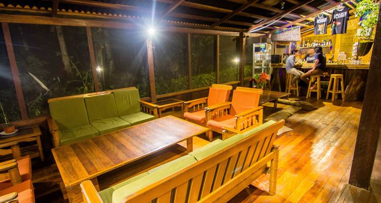 Casual wooden lounge and bar area of a jungle lodge at night with sofas, stools and two guests chatting.
