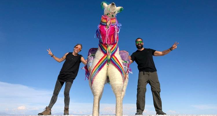 Dos personas posando junto a una escultura colorida de llama en un salar bajo un cielo azul despejado.