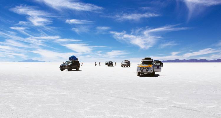 Vehículos conduciendo en una vasta planicie salina bajo un cielo azul despejado con montañas distantes.