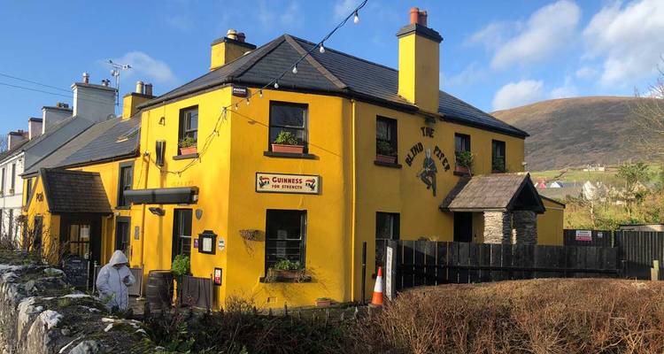 Un pub ou une auberge jaune vif avec de la signalisation, entouré de buissons et avec un décor montagneux en arrière-plan.
