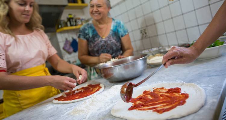 Mujeres sonrientes preparan pizzas frescas en una encimera de mármol durante una clase de cocina interactiva.